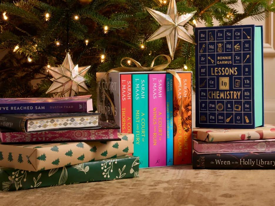 A stack of special edition books under a Christmas tree, including a Sarah J. Maas boxed set.