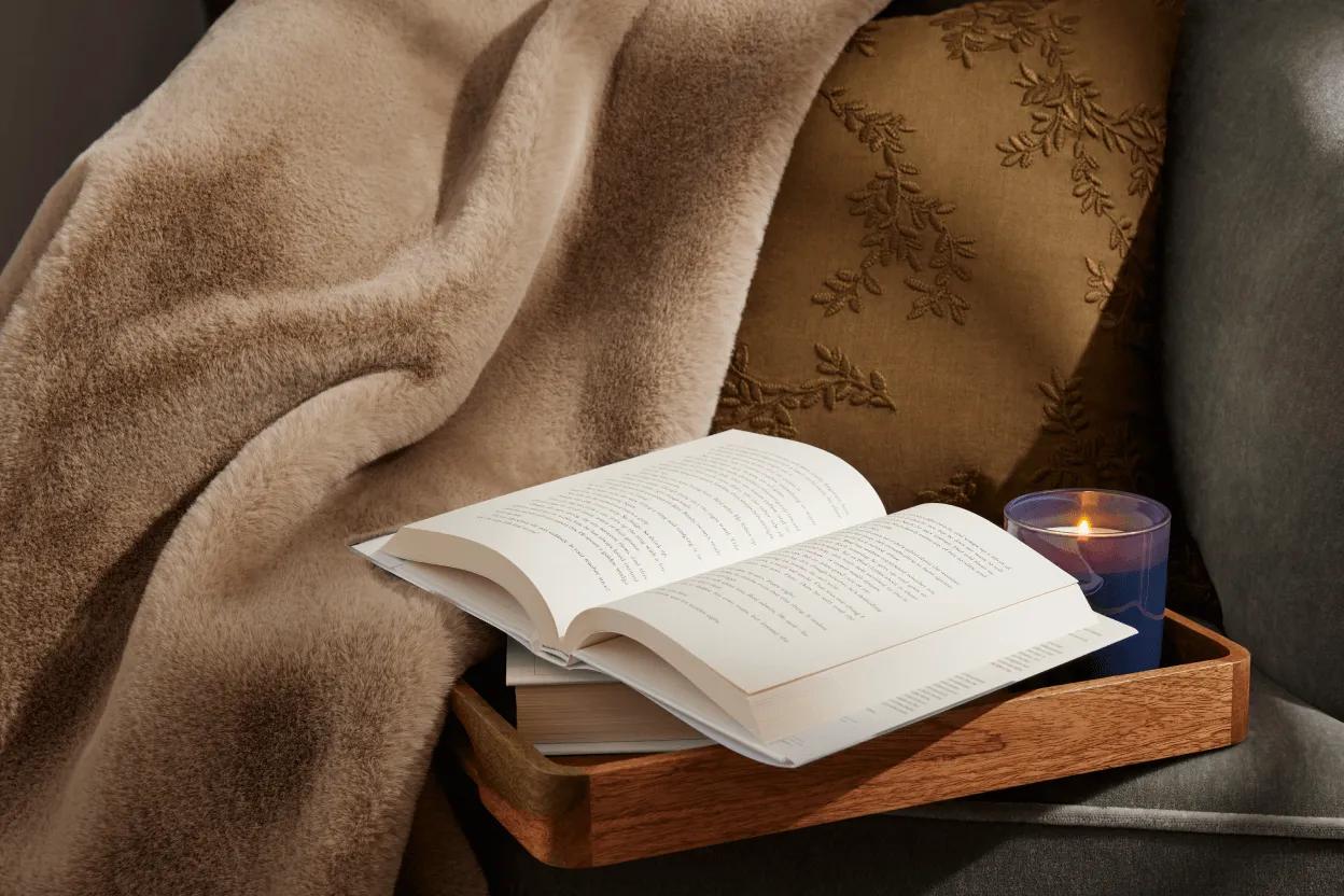 An open book on a table next to a faux fur throw and floral pillow.